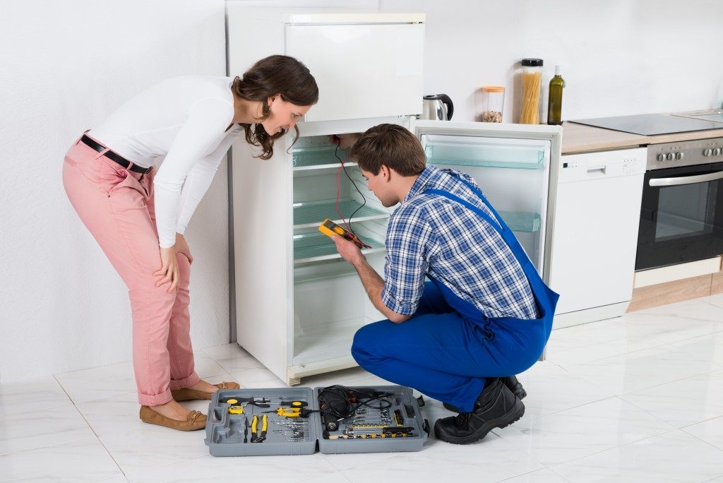 Beautiful Housewife Looking At Male Worker Repairing Refrigerator In Kitchen Room
