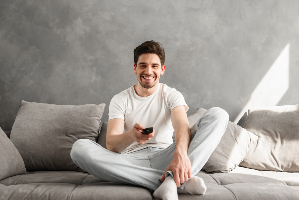 Photo of happy guy 30s in casual clothing sitting on sofa in modern flat and looking on camera with remote control in hand